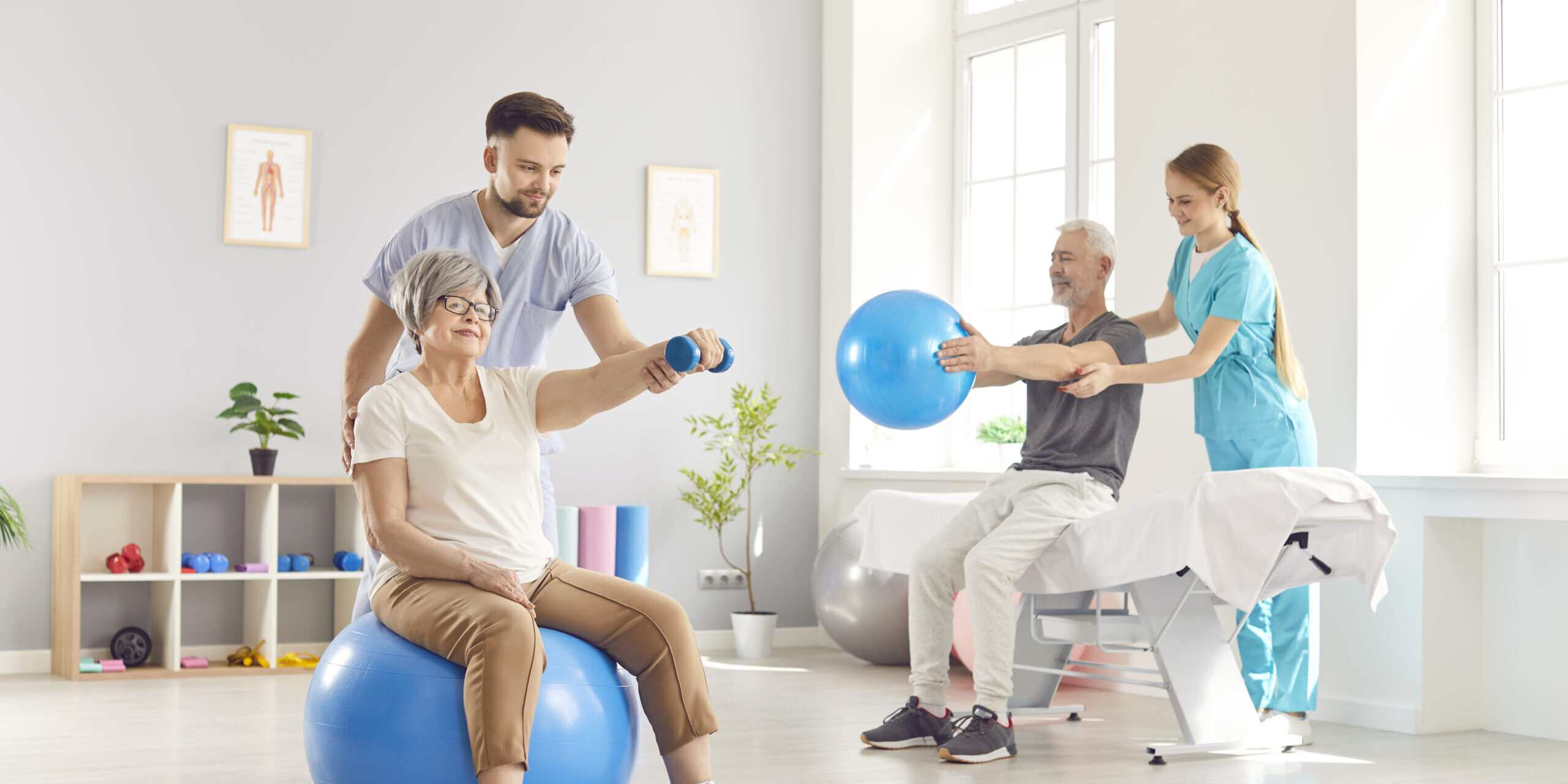 Health care workers helping mature people to do sport exercises with dumbbells and fit ball in gym.